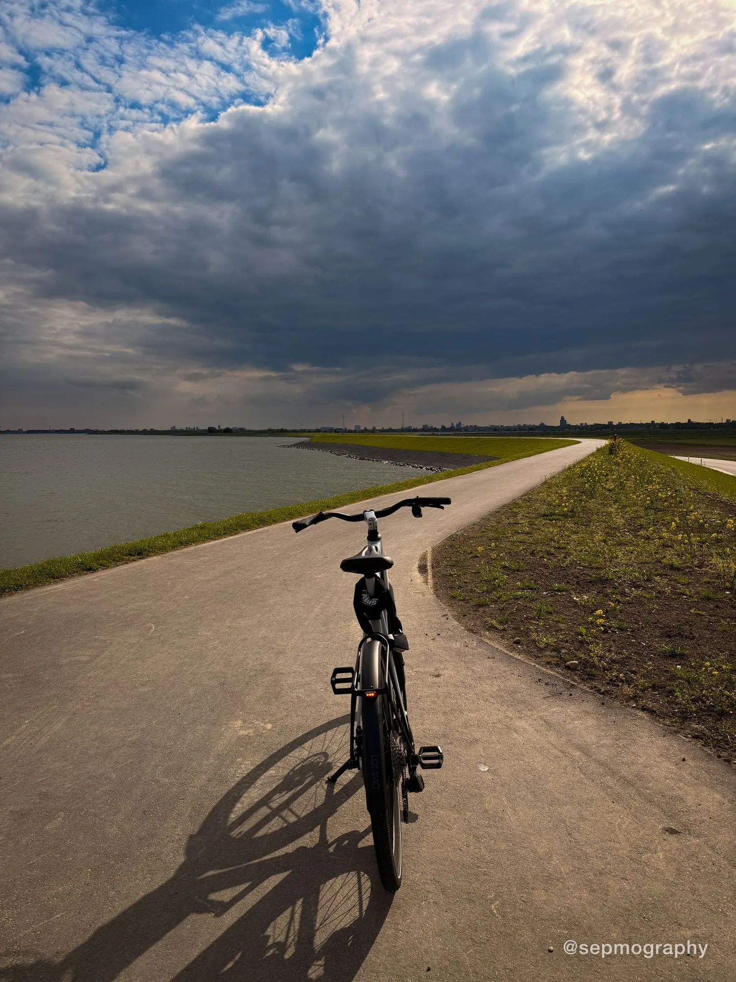 A bicycle parked on a dike path between water and grass, under a bank of storm cloud