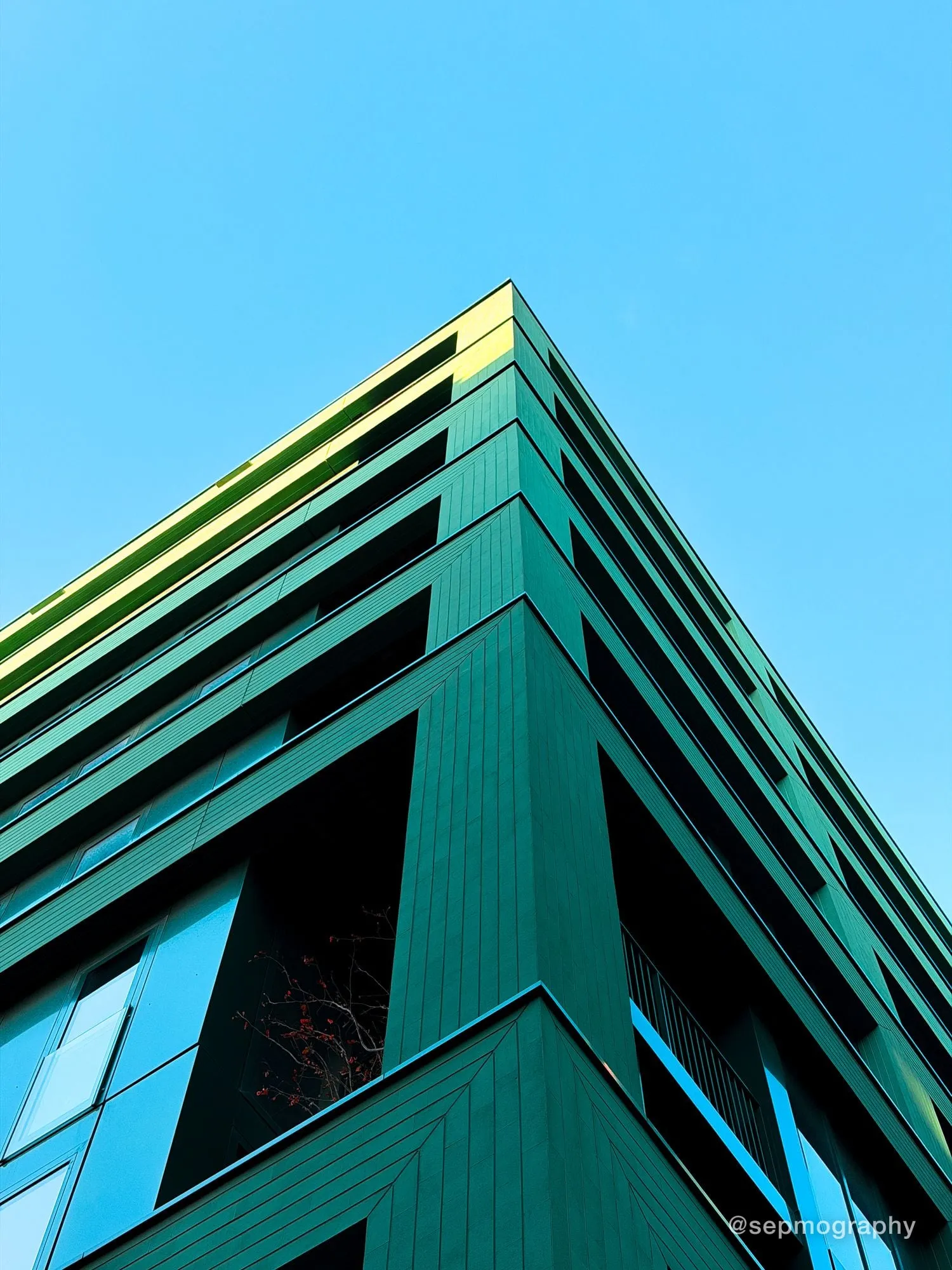The corner of a green clad building shot from below against a clear sky