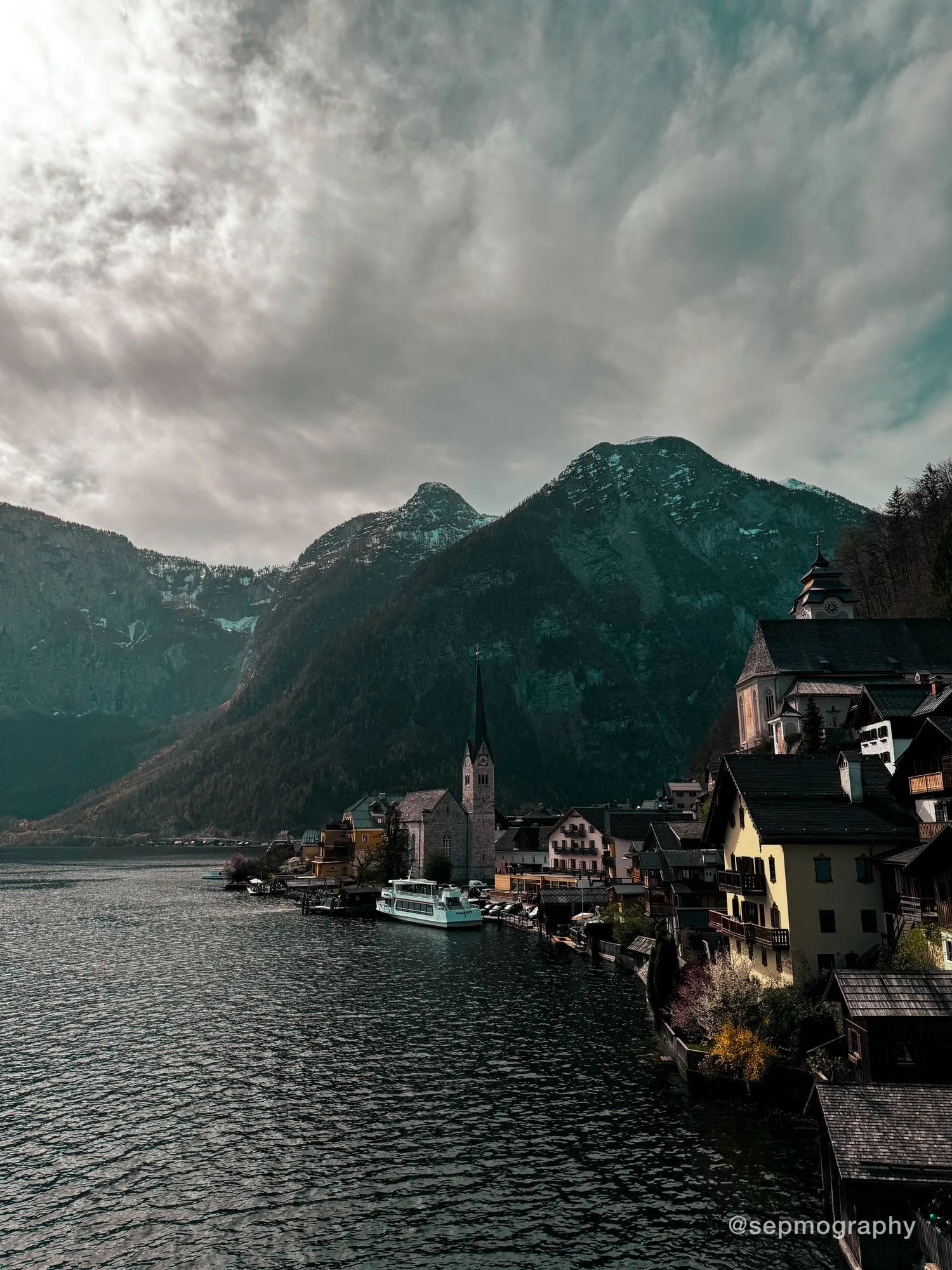 A village on the edge of a lake under overcast mountains, with a church spire at the waterline