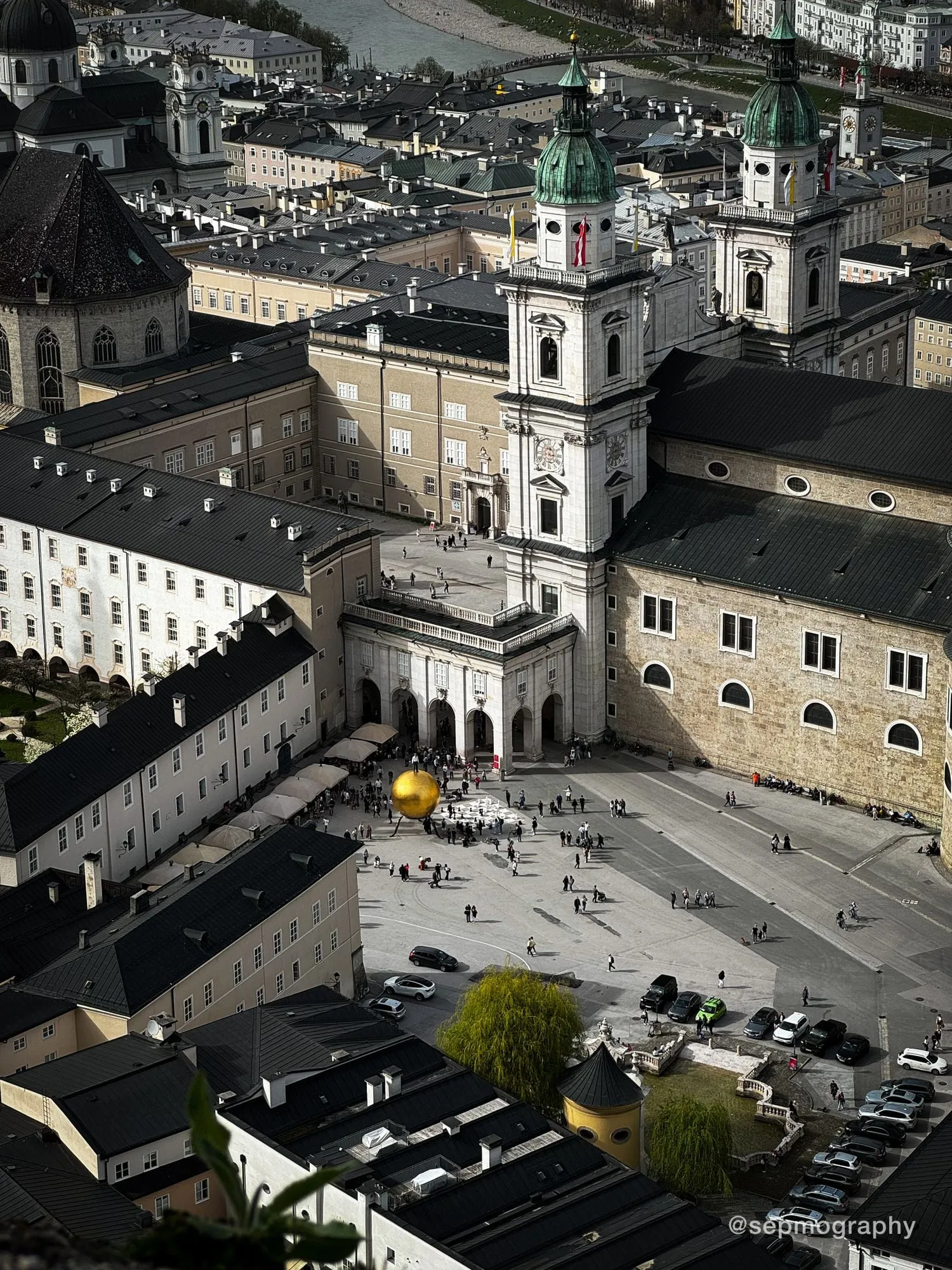 A square seen from above, with a gold sphere on a column and people crossing the stone
