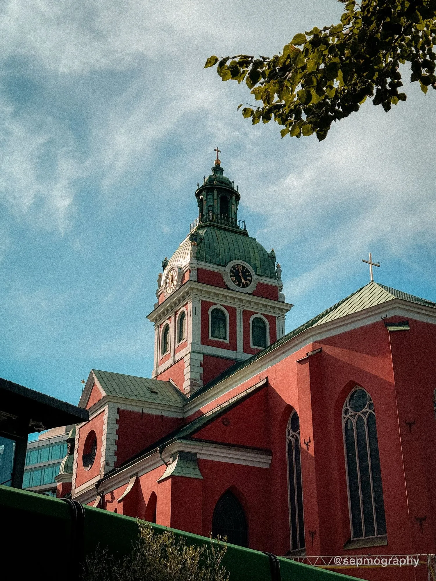 A red church with a green copper dome and a clock tower, framed by leaves